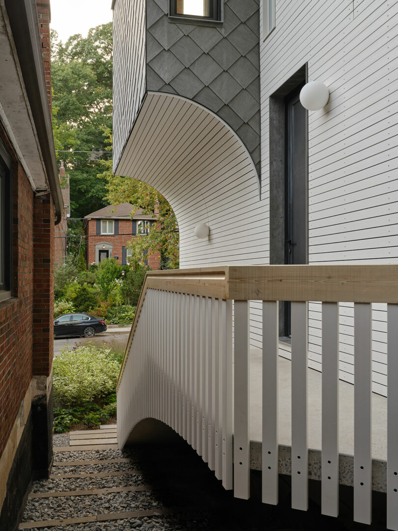A modern architectural exterior with a curved white wooden railing and contrasting grey and white siding; a tree-lined street and a black car are visible in the background.