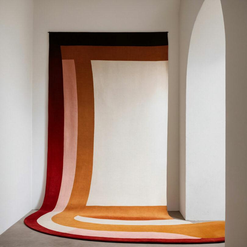 A modern, rectangular rug with curved edges in shades of red, brown, beige, and orange, draped against a white wall and extending onto a gray floor.