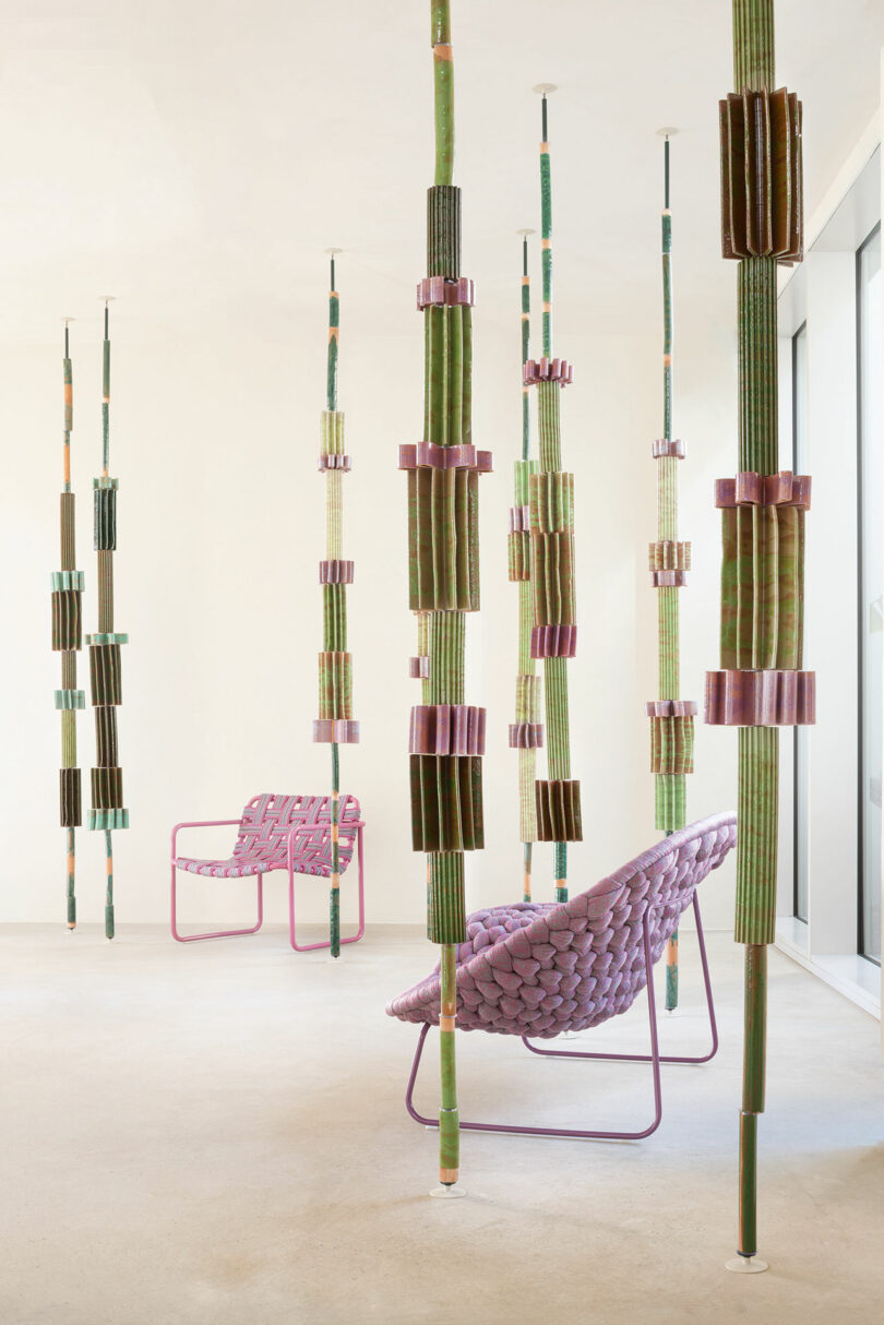 A modern interior features Stelo by Nicolò Morales for Paola Lenti, suspended columns of green and purple cylindrical elements. This is complemented by two purple woven chairs with pink frames on a minimalist beige floor.
