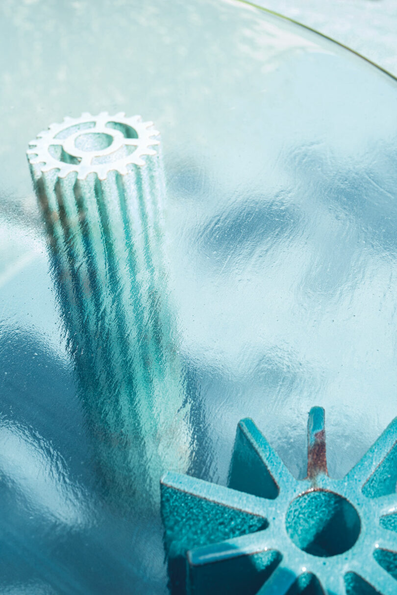 Close-up image of Helico by Nicolò Morales for Paola Lenti, featuring two textured and gear-like objects on a reflective blue surface. The objects have a weathered, rusty appearance.