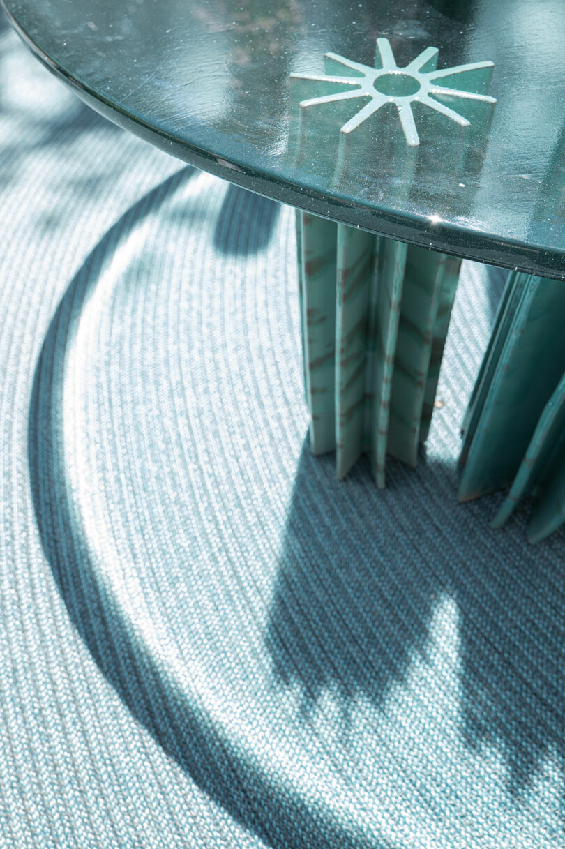 Close-up of Helico by Nicolò Morales for Paola Lenti, featuring a glass table with a sun design on the surface. This is supported by textured, geometric legs, casting shadows on a woven, light-colored floor covering.