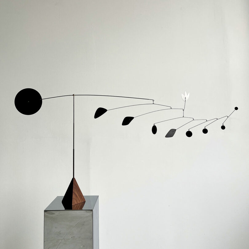 A minimalist metal mobile sculpture with various black and one white geometric shapes, mounted on a wooden and metal base, is displayed against a plain white wall.