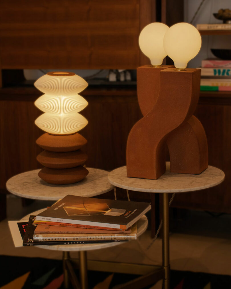Two modern table lamps with unique designs sit on round marble tables. One lamp features a wavelike pattern, while the other has two spherical bulbs.
