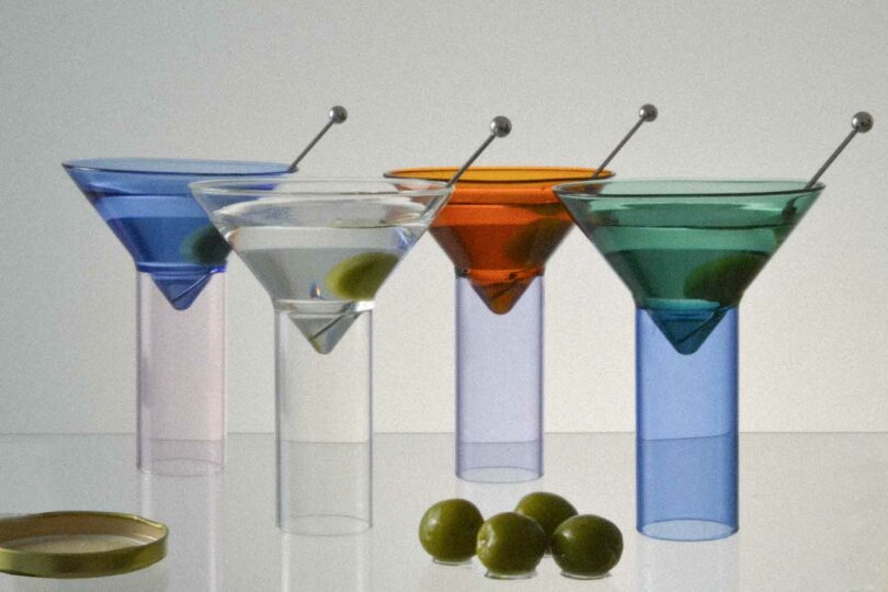 Four colorful martini glasses with different colored drinks and olives on sticks inside, arranged in a row. Three olives are placed on the surface in front of the glasses