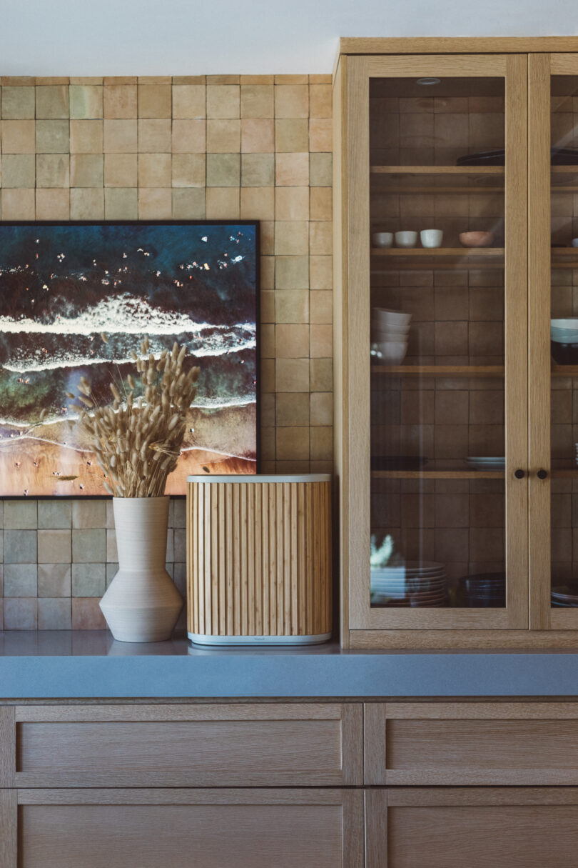 A kitchen counter with a potted plant, a basket, a modern vase, and a wooden glass cabinet. A framed abstract painting is mounted on a tiled wall in the background.