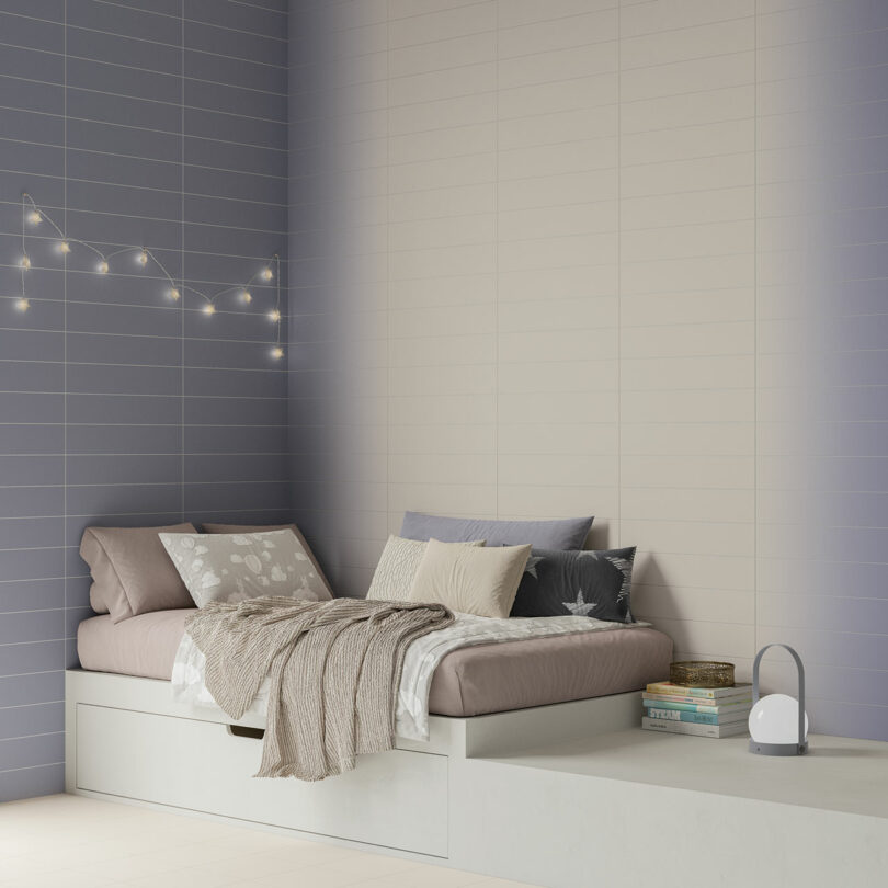 A minimalist bedroom with a bed against a two-toned wall, string lights, decorative pillows, a blanket, and a small lamp on a platform.